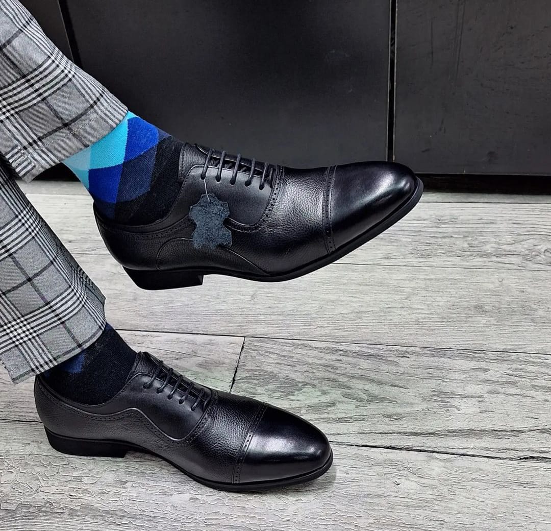 a man wearing black leather Oxford dress shoes with a lace-up cap-toe design, paired with patterned socks and tailored trousers on a clear background.